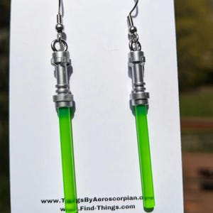 May include: A pair of silver-toned earrings with a green lightsaber design. The earrings have a detailed metallic handle and a bright green, cylindrical blade. The earrings are displayed on a white card with website information.