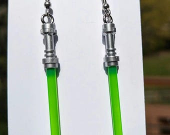 May include: A pair of silver-toned earrings with a green lightsaber design. The earrings have a detailed metallic handle and a bright green, cylindrical blade. The earrings are displayed on a white card with website information.