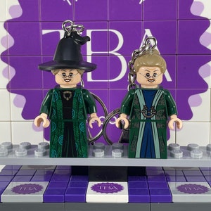 May include: Two green and blue LEGO minifigures of female characters wearing robes and hats. The minifigures are on a gray LEGO baseplate with purple LEGO bricks.