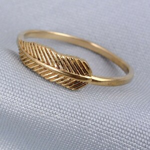 Op de afbeelding: Een goudkleurige ring met een gedetailleerd veermotief. De ring heeft een dunne band en de veer is bovenop geplaatst. De ring is gemaakt van een glanzend, metalen materiaal.