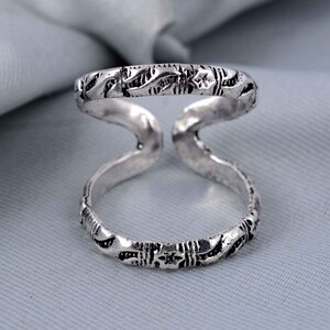 Könnte beinhalten: Ein silberfarbener Doppelbandring mit offenem Design. Der Ring weist auf den Bändern aufwendige geätzte Muster mit einem floralen oder blattartigen Motiv auf. Der Ring wird vor einem weichen, hellgrauen Hintergrund präsentiert.