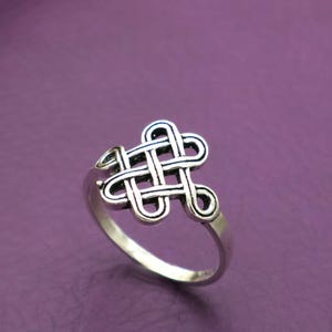 May include: A silver-toned ring with an intricate Celtic knot design. The ring features a detailed, interwoven pattern at the center, set against a purple background. The band is smooth and simple, complementing the ornate knot.