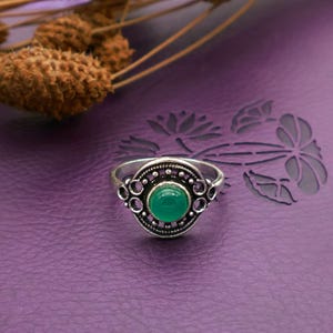 Könnte beinhalten: Ein silberfarbener Ring mit einem ovalen, hellgrünen Stein in der Mitte. Der Ring hat ein verziertes Design mit kleinen kreisförmigen Aussparungen um den Stein. Der Ring ist vor einem lila Hintergrund platziert.
