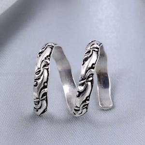 May include: A silver-toned metal ring featuring a continuous spiral design. The surface of the band is decorated with an embossed, repeating floral or vine-like pattern that has been darkened in the recessed areas to highlight the intricate details.