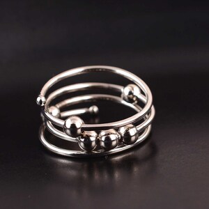 Op de afbeelding: Een zilverkleurige metalen ring met een spiraalvormig ontwerp met meerdere windingen. De band bestaat uit drie overlappende cirkelvormige lussen, met verschillende kleine, gladde, bolvormige metalen kralen aan de draad die langs de band kunnen schuiven.