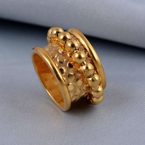 Könnte beinhalten: Ein goldfarbener Ring mit einem strukturierten Band mit einem Punktmuster und einer Reihe kleiner, runder Perlen. Der Ring hat eine polierte Oberfläche und ist ein Schmuckstück.