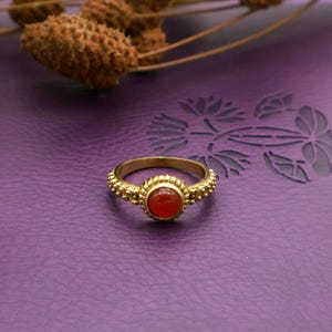Puede incluir: Un anillo dorado con una piedra preciosa roja redonda en el centro. El anillo tiene una banda texturizada con pequeños detalles en relieve. El anillo está sobre un fondo morado, con tallos de plantas secas en el fondo.