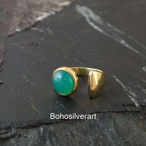 Puede incluir: Un anillo abierto de tono dorado con una piedra cabujón redonda, lisa y de color verde vibrante en un extremo y un acento de metal sólido pulido de tono dorado en el otro. El texto "Bohosilverart" está impreso en blanco en la parte inferior.