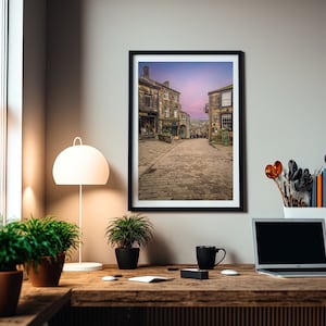 May include: A framed photograph of a cobblestone street in a small town with buildings on either side. The sky is a soft pink and purple color.