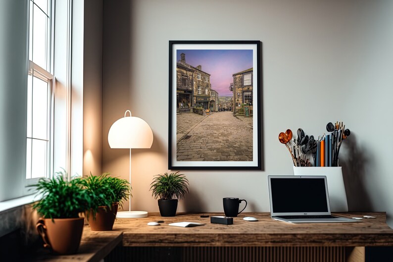May include: A framed photograph of a cobblestone street in a small town with buildings on either side. The sky is a soft pink and purple color.