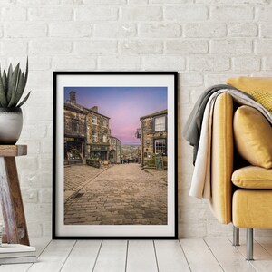 May include: A framed photograph of a cobblestone street in a historic town. The street is lined with old stone buildings, and the sky is a soft purple color.