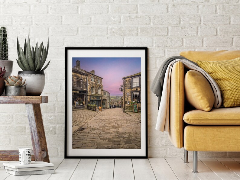 May include: A framed photograph of a cobblestone street in a historic town. The street is lined with old stone buildings, and the sky is a soft purple color.