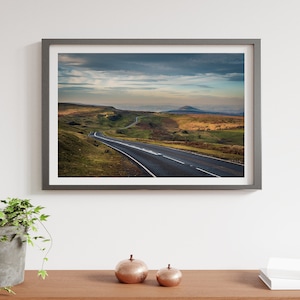 May include: A framed photograph of a winding road leading through a green valley with a mountain in the distance. The sky is cloudy and the sun is setting.