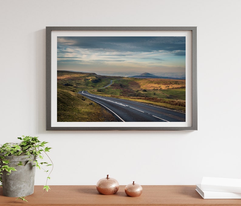 May include: A framed photograph of a winding road leading through a green valley with a mountain in the distance. The sky is cloudy and the sun is setting.