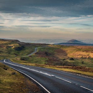 May include: A winding paved road with white lane markings leads through a green grassy valley with a distant mountain range in the background. The sky is a light blue with white clouds.