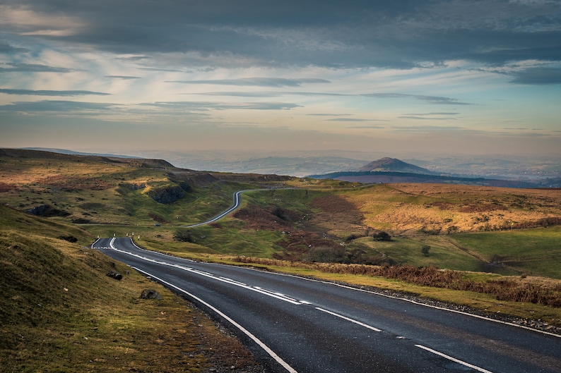 May include: A winding paved road with white lane markings leads through a green grassy valley with a distant mountain range in the background. The sky is a light blue with white clouds.