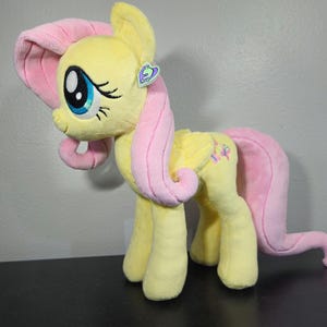 May include: A yellow and pink plush pony toy. The pony features a yellow body, pink mane and tail, and large blue eyes. It has a tag on its ear. The toy is standing on a black surface.