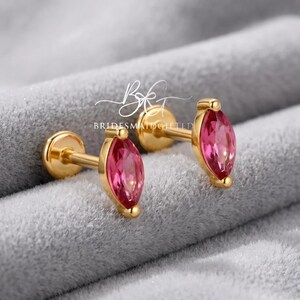 Pode incluir: Um par de brincos de pino dourados com pedras preciosas cor-de-rosa em corte marquise. Cada brinco tem um fecho circular plano e um pino. O texto BRIDESMAIDGIFTED é visível no centro. Os brincos têm um acabamento dourado polido e pedras.