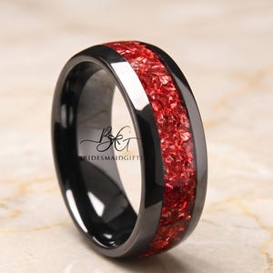 Puede incluir: Un anillo negro con una banda de piedra roja triturada. El anillo tiene un acabado brillante y una forma lisa y redondeada. El texto "BRIDESMAIDGIFT" es visible en el anillo.
