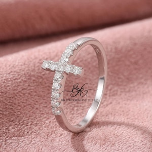 Puede incluir: Un anillo plateado con un diseño de cruz. La cruz está incrustada con pequeñas piedras preciosas transparentes. La banda del anillo también tiene piedras preciosas. El anillo se muestra sobre una superficie rosa.