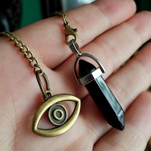 May include: A necklace featuring a polished black stone point pendant set in a silver-toned metal cap, attached to a bronze-toned chain with an eye-shaped charm. The pendant hangs from a lobster claw clasp, showcasing a contrast between the dark stone and the mixed metal finishes of the chain and eye charm.