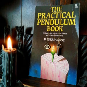 May include: A book titled The Practical Pendulum Book by D. Jurriaanse featuring two metal chains with pendants. One pendant is a gold-toned eye symbol, and the other is a black stone point. A lit candle stands in the foreground.