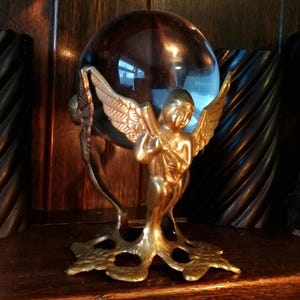 May include: A decorative golden angel figurine holding a dark brown crystal ball. The angel has detailed wings and is mounted on a gold-coloured base. The crystal ball reflects light, creating a captivating display.