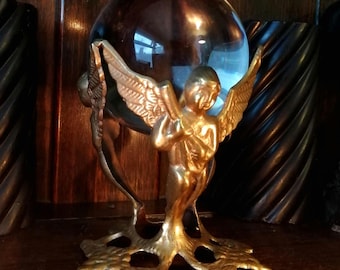 May include: A decorative golden angel figurine holding a dark brown crystal ball. The angel has detailed wings and is mounted on a gold-colored base. The crystal ball reflects light, creating a captivating display.