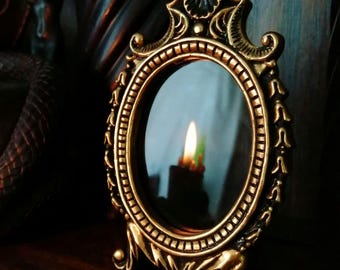 May include: Ornate, antique-style oval mirror with a gold-colored frame. The mirror reflects a candle flame, creating a warm glow. The frame features decorative details and rests on small feet. A vintage-inspired decorative piece.