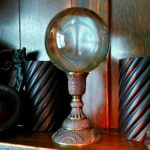 May include: A clear glass gazing ball with iridescent reflections sits atop an ornate, antique-style brass stand. The stand features detailed patterns and a wide base. The background includes dark wooden elements and decorative accents.