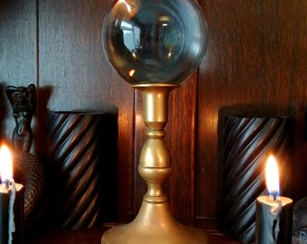 May include: A clear crystal ball sits atop a gold-colored metal stand. The stand has a wide base and a decorative, bulbous stem. The background includes dark wooden panels and lit candles, creating a mystical ambiance.