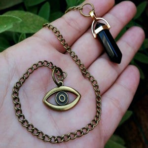 May include: A bronze-toned metal chain necklace featuring two distinct pendants. One pendant is a smooth, black, hexagonal crystal point capped in bronze metal. The second pendant is a bronze-toned eye symbol with a circular center detail.