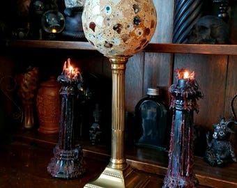 May include: A gold metal lamp base with a round, white and brown mosaic glass shade. The lamp is on a wooden shelf with two black candle holders with red wax dripping down the sides.