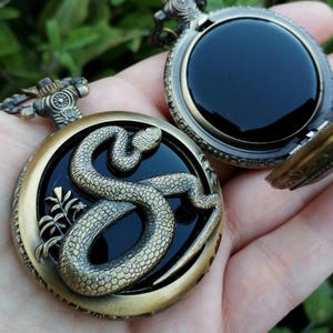 May include: Vintage bronze pocket watch featuring a detailed snake design on a black face. The watch is open, revealing the black interior. It has a decorative chain attachment and a textured finish.