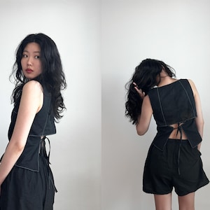 May include: A black sleeveless top featuring a square neckline, white contrast stitching along the edges, and a tie-back closure at the waist, paired with black shorts that have a scalloped hemline detail along the bottom edge of the fabric.