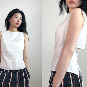 May include: A sleeveless white top with a boat neckline and a vertical seam down the center front. The garment features a cropped back panel and a tie closure at the waist, paired with black trousers featuring vertical white and tan stripes.