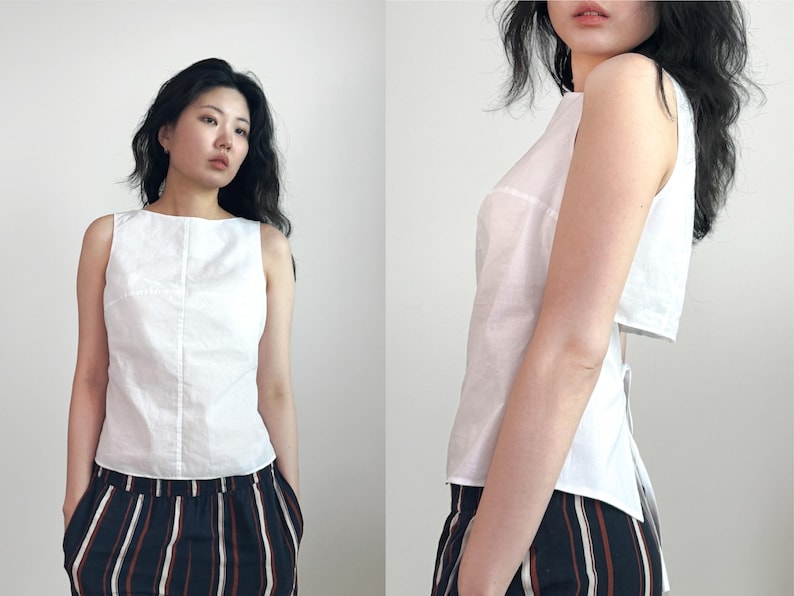 May include: A sleeveless white top with a boat neckline and a vertical seam down the center front. The garment features a cropped back panel and a tie closure at the waist, paired with black trousers featuring vertical white and tan stripes.