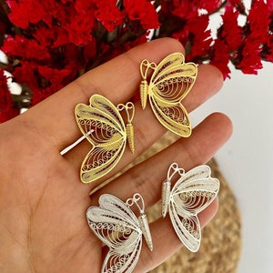 May include: Close-up of three filigree butterfly pendants in gold and silver. The butterfly wings have intricate openwork designs. The pendants are displayed against a background of red flowers.