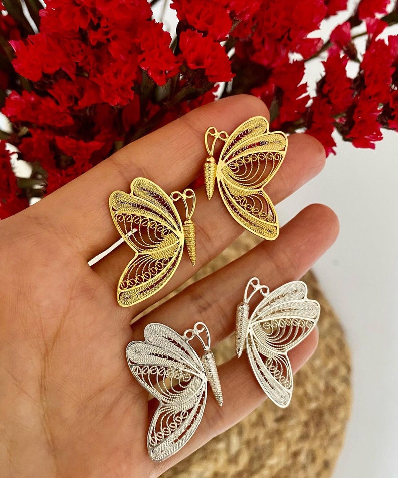 May include: Close-up of three filigree butterfly pendants in gold and silver. The butterfly wings have intricate openwork designs. The pendants are displayed against a background of red flowers.