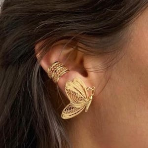 May include: Gold-colored butterfly earring and ear cuff. The butterfly earring features intricate filigree detailing, with delicate wings and a detailed body. The ear cuff has a geometric design. The jewelry is worn on the ear.