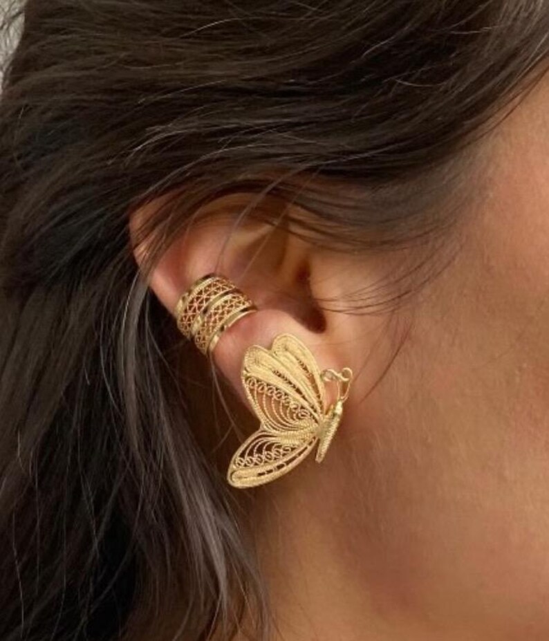 May include: Gold-colored butterfly earring and ear cuff. The butterfly earring features intricate filigree detailing, with delicate wings and a detailed body. The ear cuff has a geometric design. The jewelry is worn on the ear.