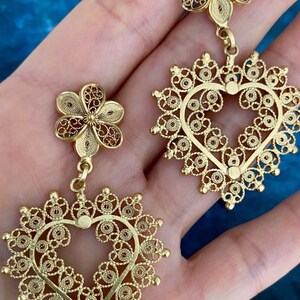 May include: Gold-colored filigree earrings. Each earring features a flower-shaped stud and a heart-shaped pendant. The intricate design showcases delicate patterns and a warm, metallic hue, perfect for adding a touch of elegance.