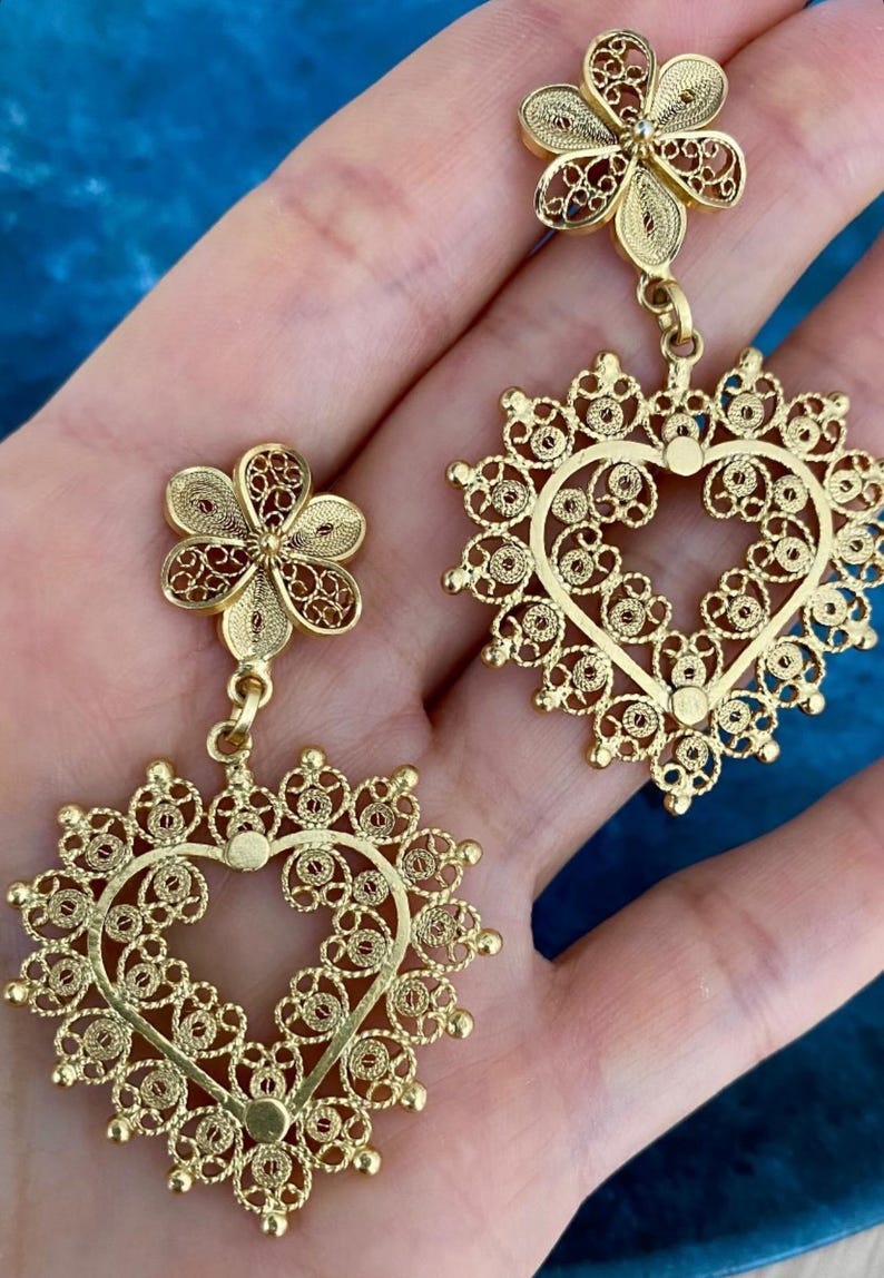 May include: Gold-colored filigree earrings. Each earring features a flower-shaped stud and a heart-shaped pendant. The intricate design showcases delicate patterns and a warm, metallic hue, perfect for adding a touch of elegance.