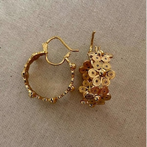 May include: A pair of gold-colored hoop earrings. One earring features a floral design, while the other has a dotted pattern along the hoop. The earrings are displayed on a neutral-colored fabric background.