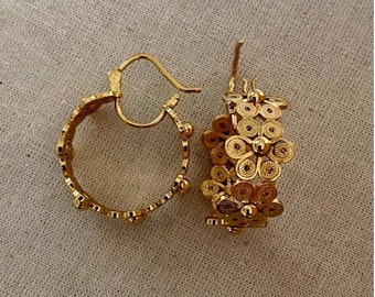 May include: A pair of gold-colored hoop earrings. One earring features a floral design, while the other has a dotted pattern along the hoop. The earrings are displayed on a neutral-colored fabric background.