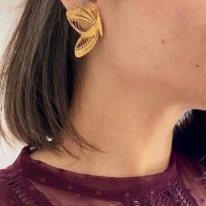 May include: A gold-colored butterfly earring is worn on the ear. The earring is a detailed filigree design, with openwork patterns creating the shape of the butterfly. The earring is a statement piece, adding a touch of elegance.