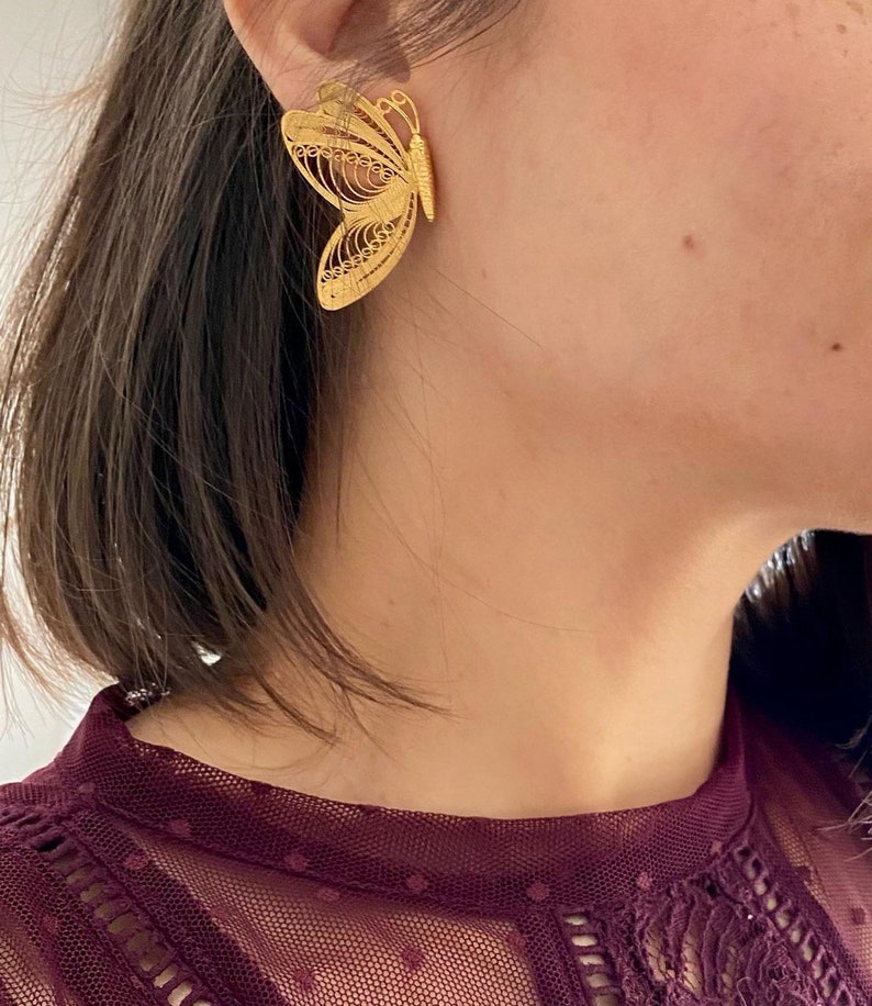 May include: A gold-colored butterfly earring is worn on the ear. The earring is a detailed filigree design, with openwork patterns creating the shape of the butterfly. The earring is a statement piece, adding a touch of elegance.
