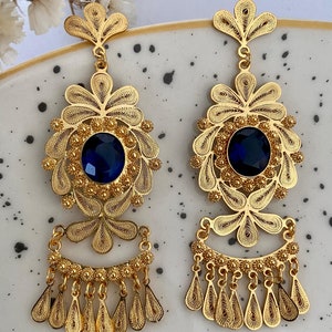 May include: A pair of gold filigree earrings with blue gemstone accents. The earrings feature a large, round, blue gemstone surrounded by intricate gold filigree designs. The earrings have a dangling, tassel-like design with multiple gold drops.