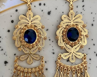 May include: A pair of gold filigree earrings with blue gemstone accents. The earrings feature a large, round, blue gemstone surrounded by intricate gold filigree designs. The earrings have a dangling, tassel-like design with multiple gold drops.