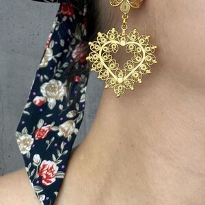 May include: Gold-toned filigree heart-shaped earrings with a floral accent. The earrings feature intricate openwork designs and a delicate, lacy appearance. A dark blue floral scarf is visible in the background.